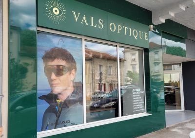 Façade du magasin Vals Optique avec enseigne verte, logo œil stylisé et vitrine avec affiche publicitaire de lunettes de sport