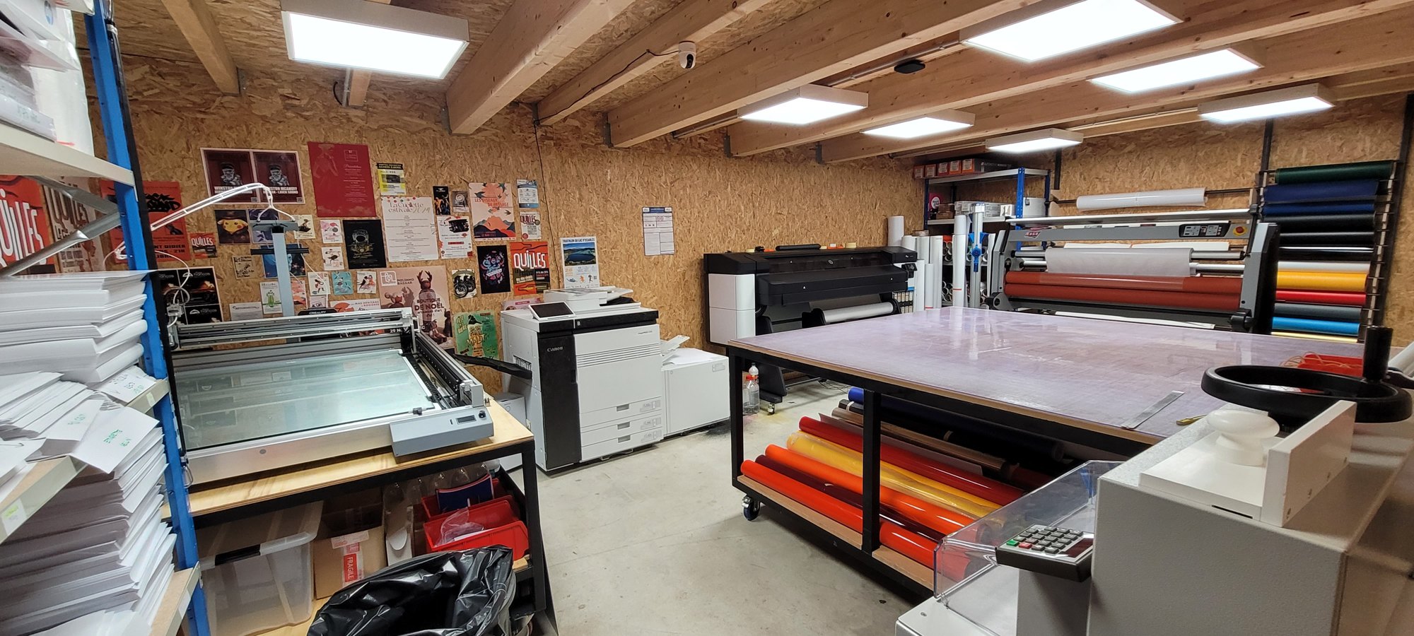 Atelier d'impression avec un traceur grand format, une table de façonnage et un rack de rouleaux de vinyle de couleur rangés sous le plateau
