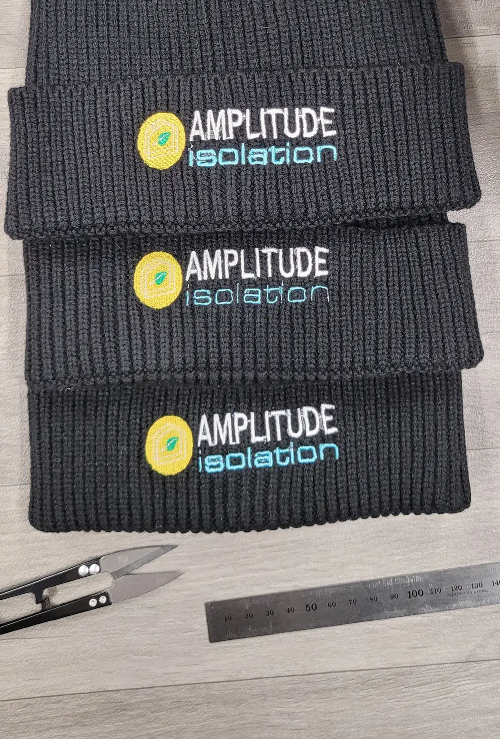 Trois bonnets anthracite brodés du logo Amplitude Isolation en blanc, jaune et cyan, posés près de ciseaux de brodeur et d'une règle métallique