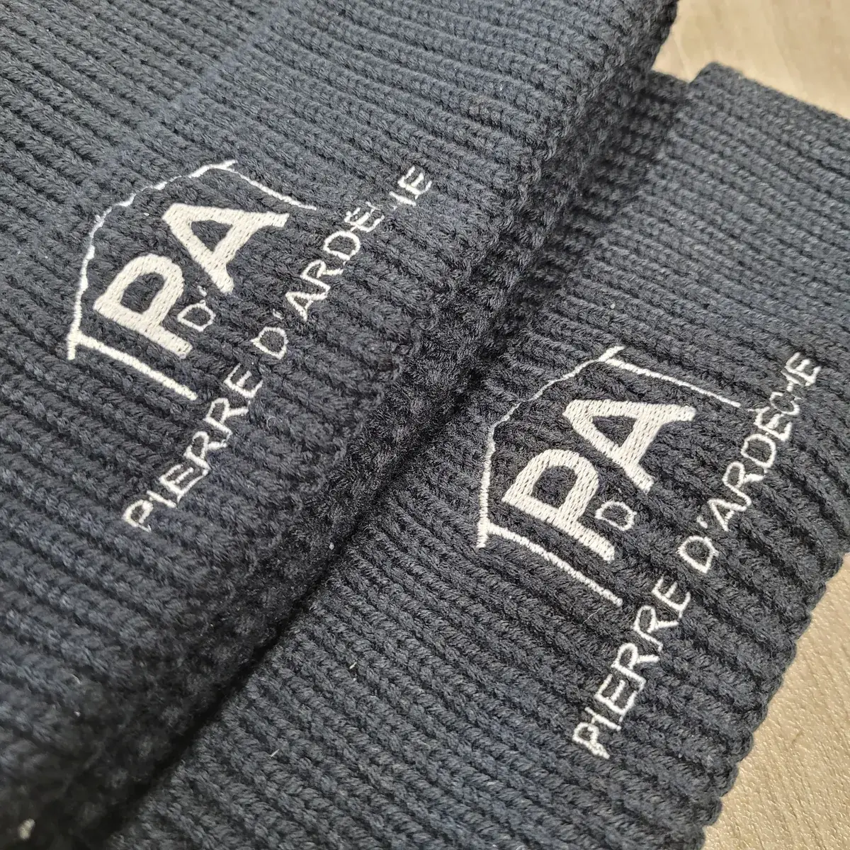 Gros plan sur la broderie en fil blanc du logo Pierre d'Ardèche, sur le revers côtelé d'un bonnet bleu marine