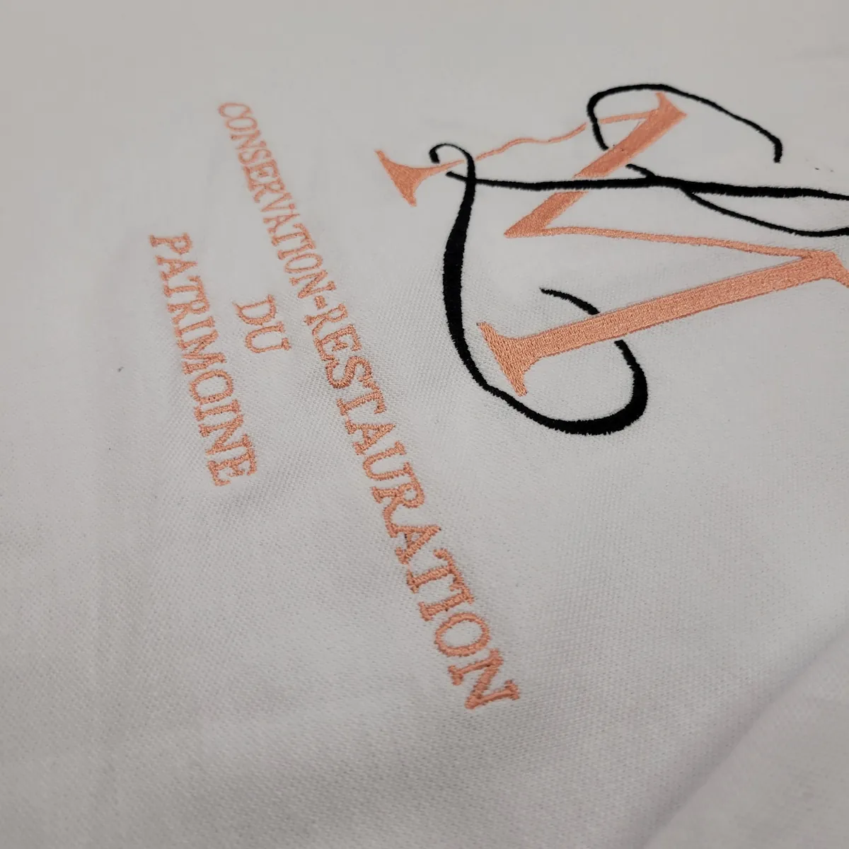 Polo blanc brodé d'un monogramme en fil rose et noir accompagné de la mention Conservation-Restauration du Patrimoine