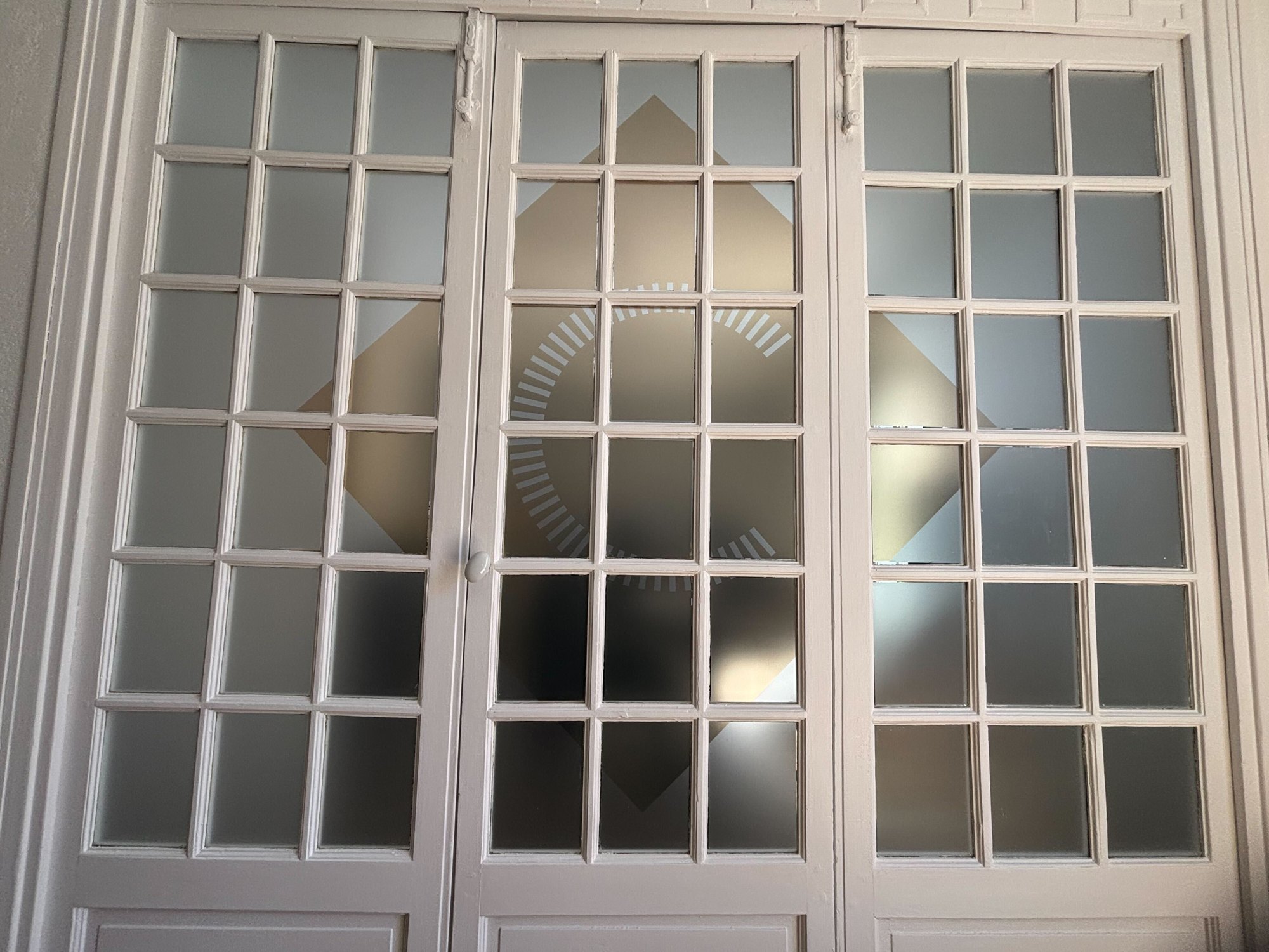 Porte-fenêtre blanche à petits carreaux dont chaque carreau est couvert de film dépoli, l'ensemble composant un motif géométrique de losange et d'arc de cercle sur les trois vantaux