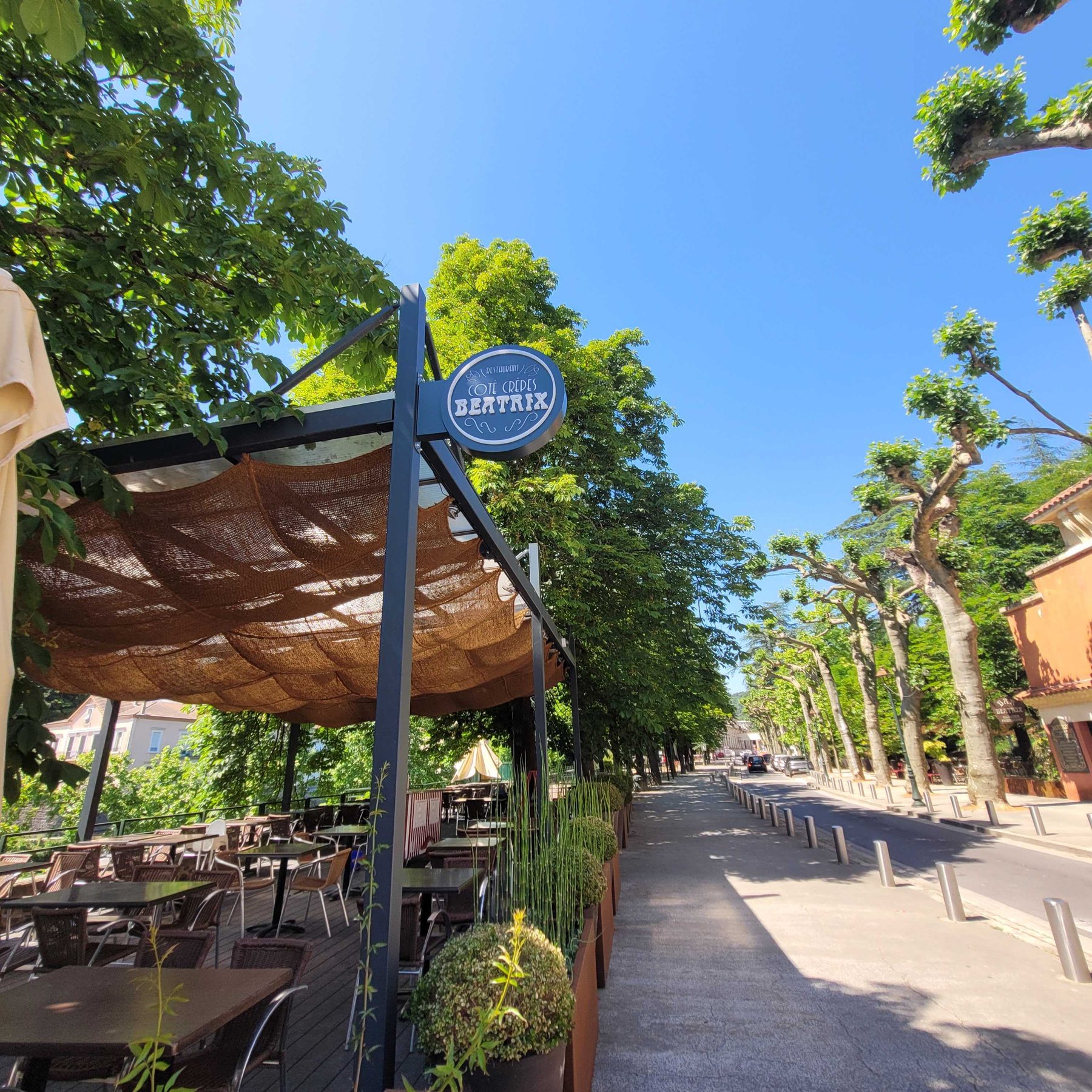 Enseigne drapeau ronde du restaurant Côte Crêpes Beatrix, suspendue en potence au-dessus de la terrasse, sous les platanes