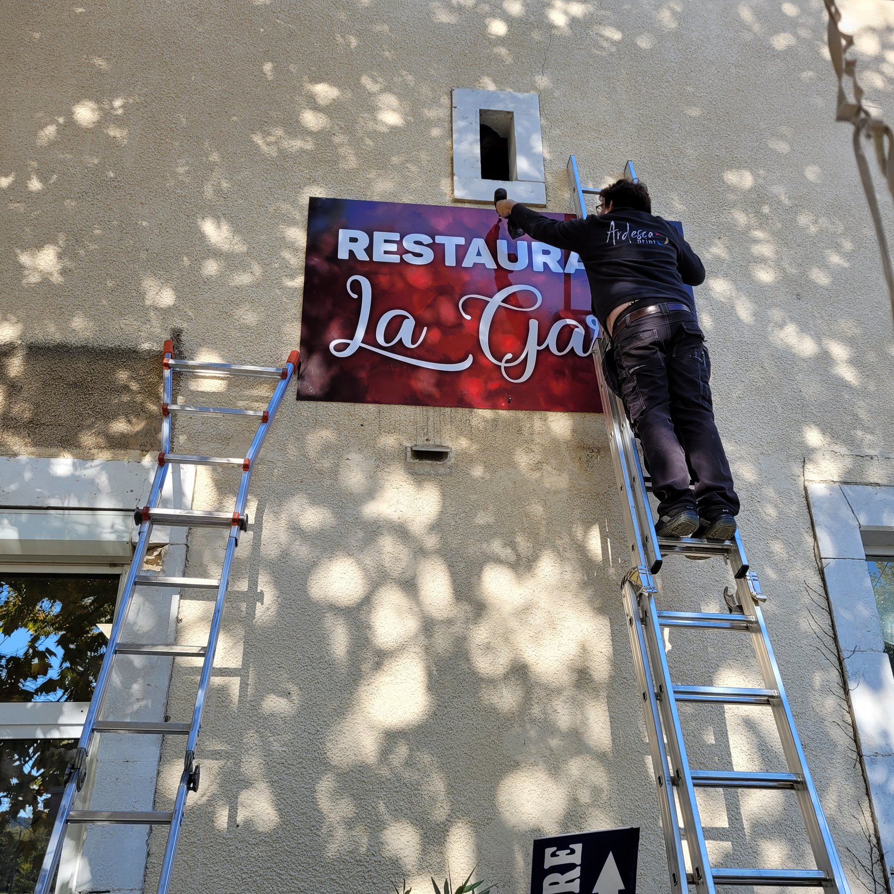 Poseur Ardesca Print sur une échelle, en train de fixer le panneau d'enseigne du restaurant La Gare sur une façade