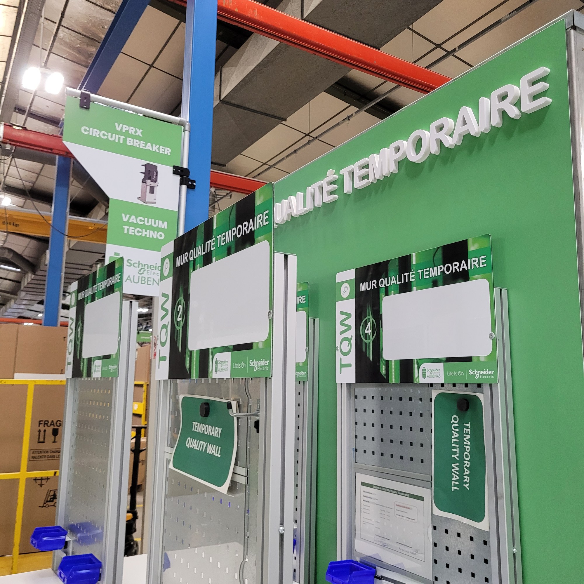 Lettres relief blanches Qualité temporaire posées sur un mur vert, avec panneaux de signalétique suspendus dans un atelier industriel