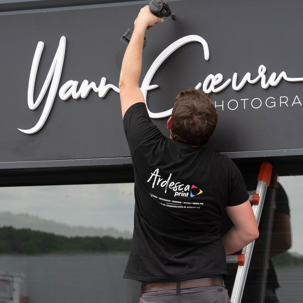 Poseur Ardesca Print sur une échelle, visseuse en main, fixant les lettres relief blanches de l’enseigne Yann Cœuru Photographe sur un bandeau noir