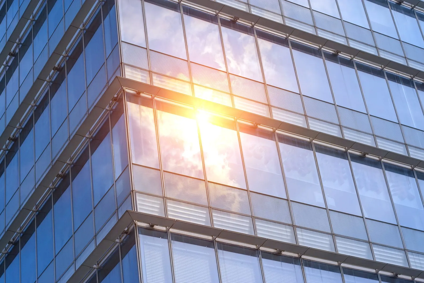 Façade d'immeuble de bureaux entièrement vitrée, le soleil se reflétant sur les panneaux, stores intérieurs baissés derrière le vitrage.