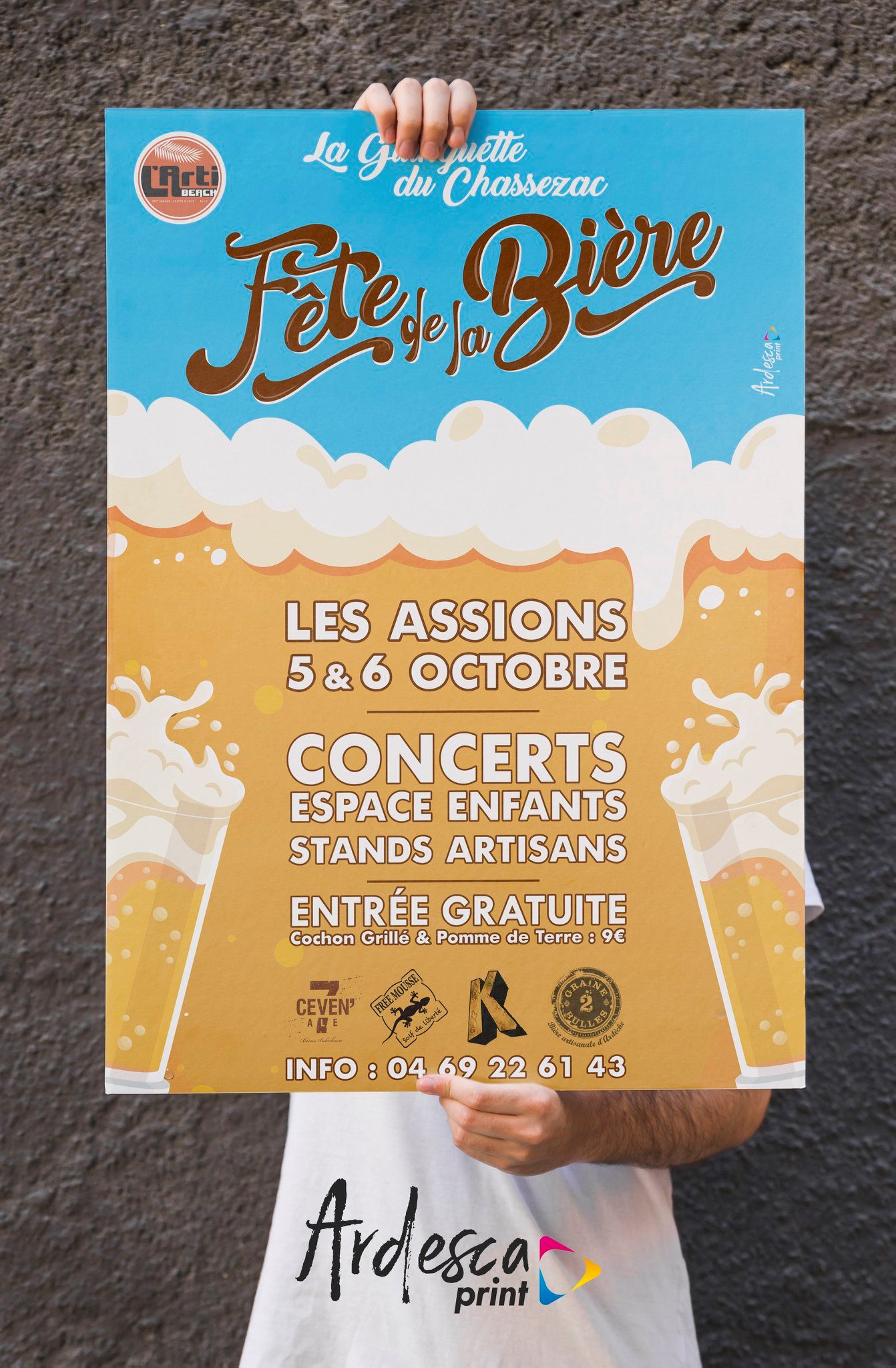 Affiche imprimée de la Fête de la Bière de La Guinguette du Chassezac aux Assions les 5 et 6 octobre, tenue à bout de bras devant un mur de pierre