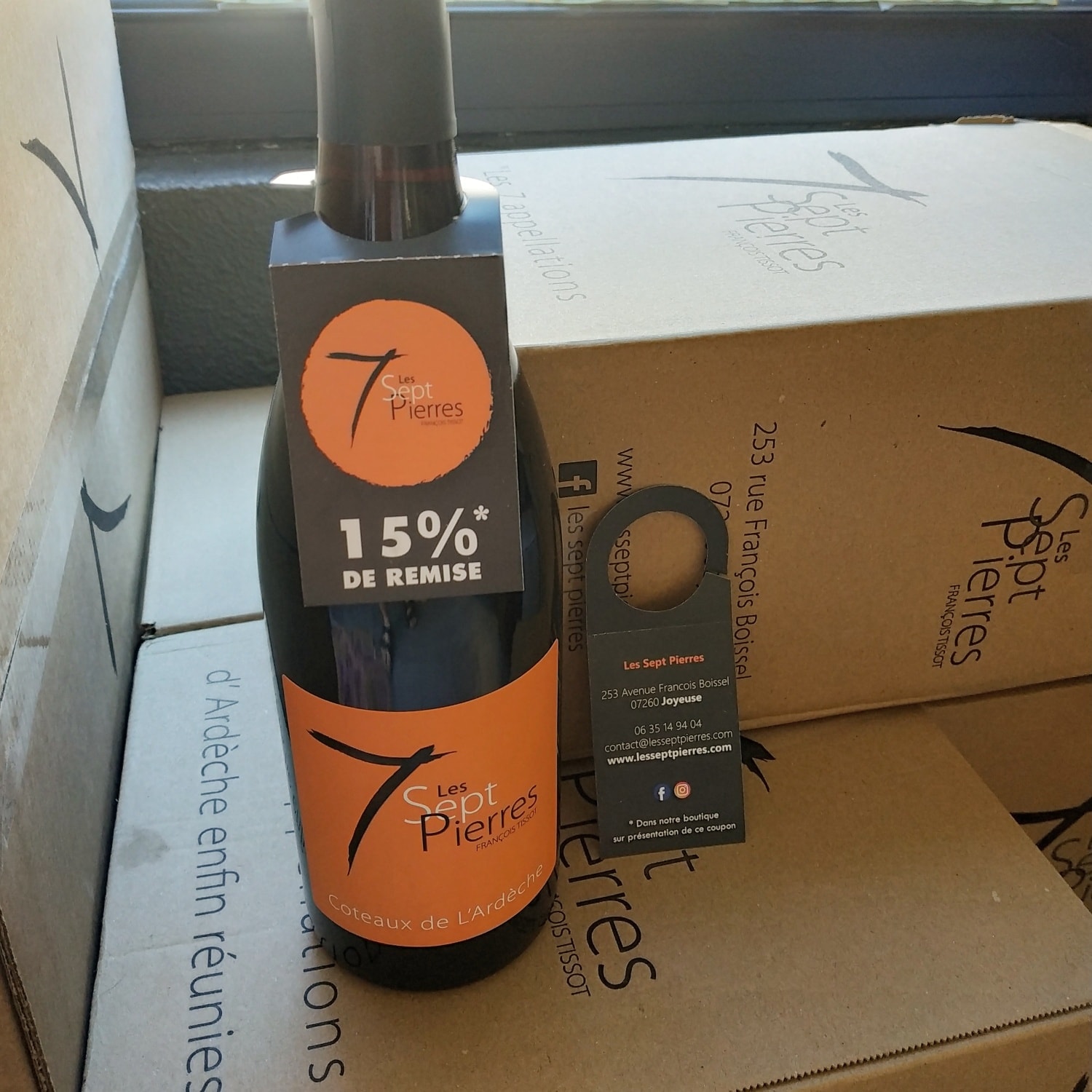 Collerette de bouteille et coupon de remise Les Sept Pierres, posés sur des cartons de vin de Joyeuse