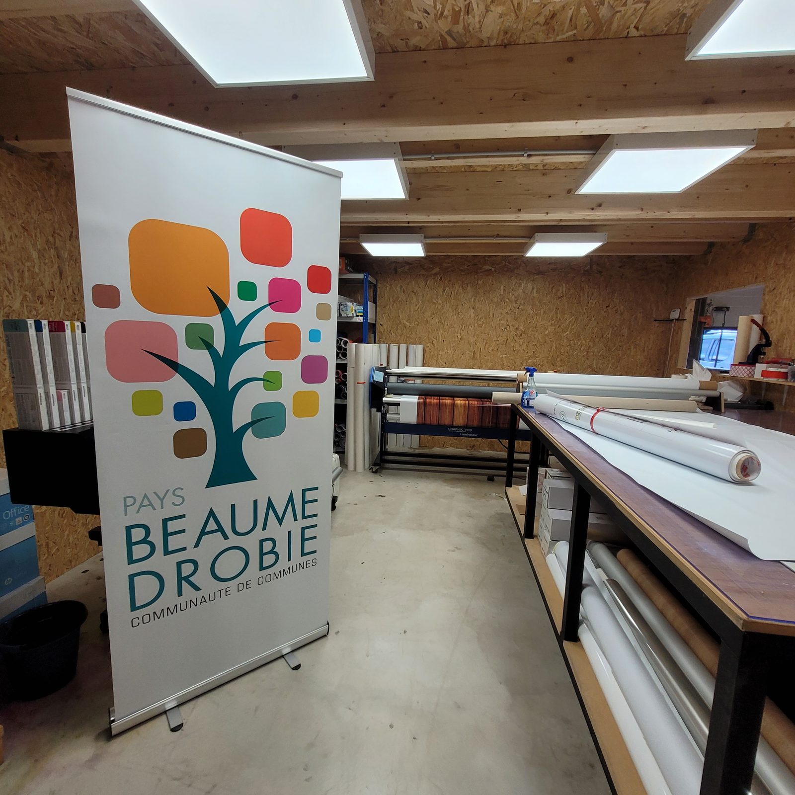 Roll-up de la communauté de communes Pays Beaume-Drobie monté dans l’atelier Ardesca, devant la plastifieuse et les rouleaux de média