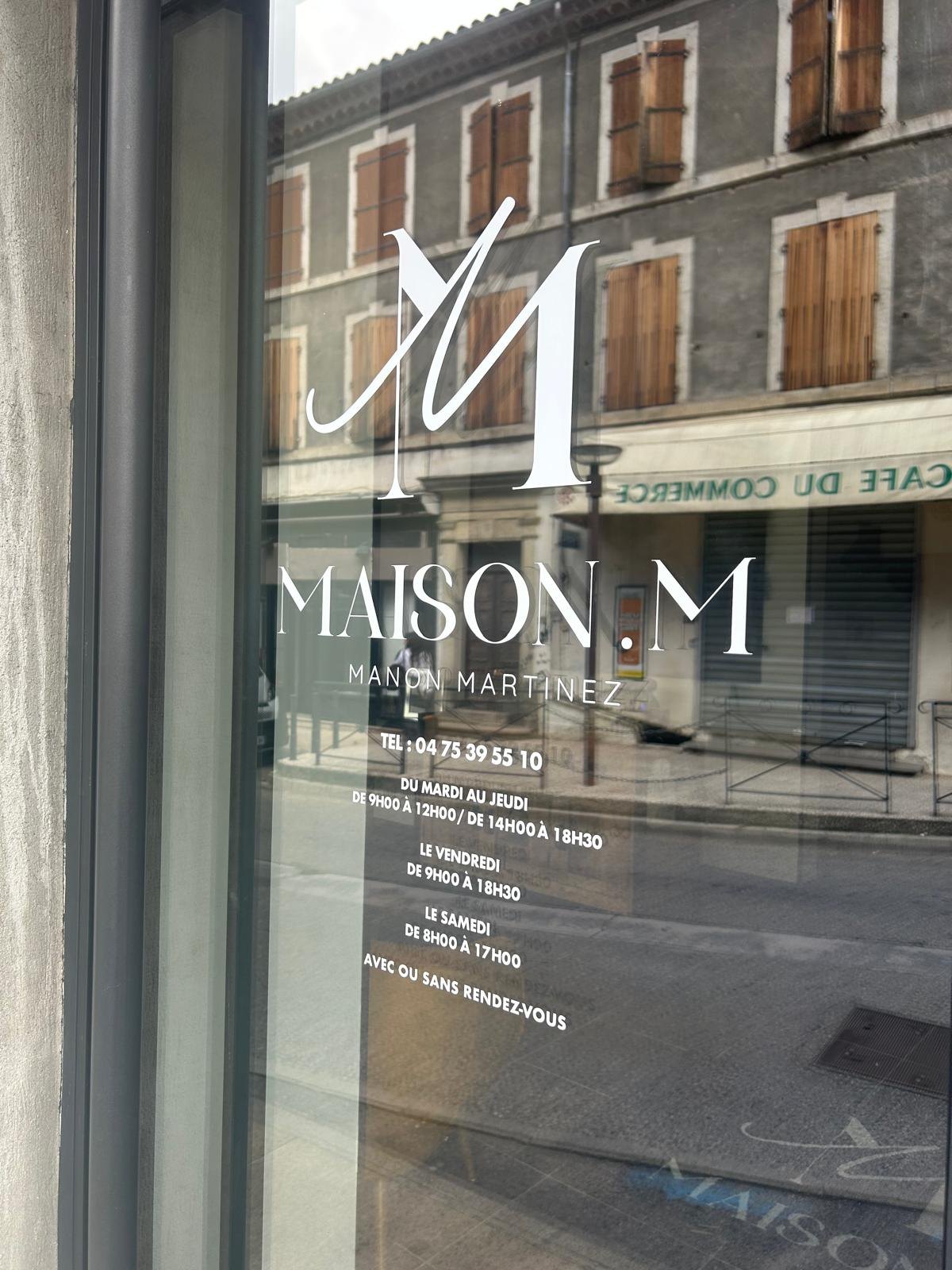Vitrine d'un salon portant en lettrage adhésif blanc un monogramme, le nom de l'enseigne, le numéro de téléphone et le détail des horaires d'ouverture