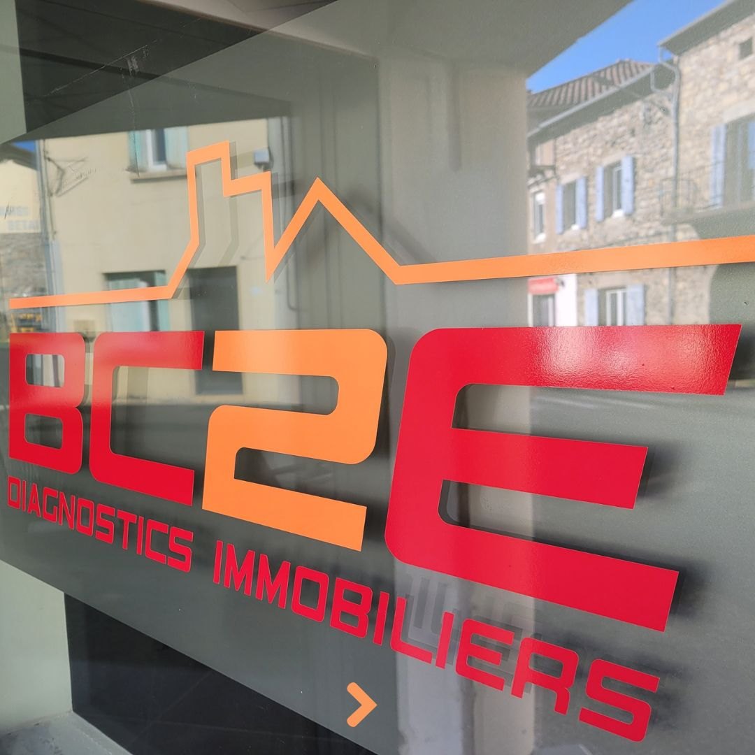 Gros plan sur un logo d'entreprise de diagnostics immobiliers découpé dans de l'adhésif rouge et orange et posé sur un vitrage, avec le tracé d'une ligne de toits au-dessus du nom