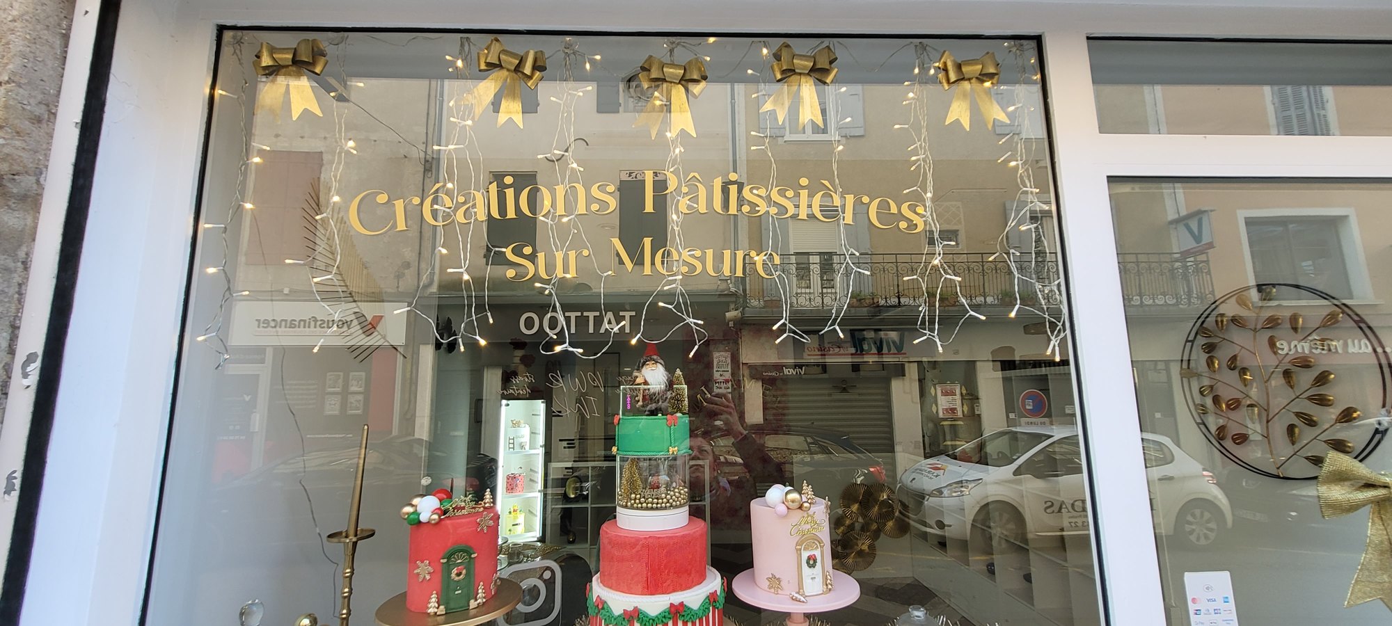 Vitrine de pâtisserie décorée pour les fêtes, dont le vitrage porte en grandes lettres adhésives dorées la mention Créations Pâtissières Sur Mesure