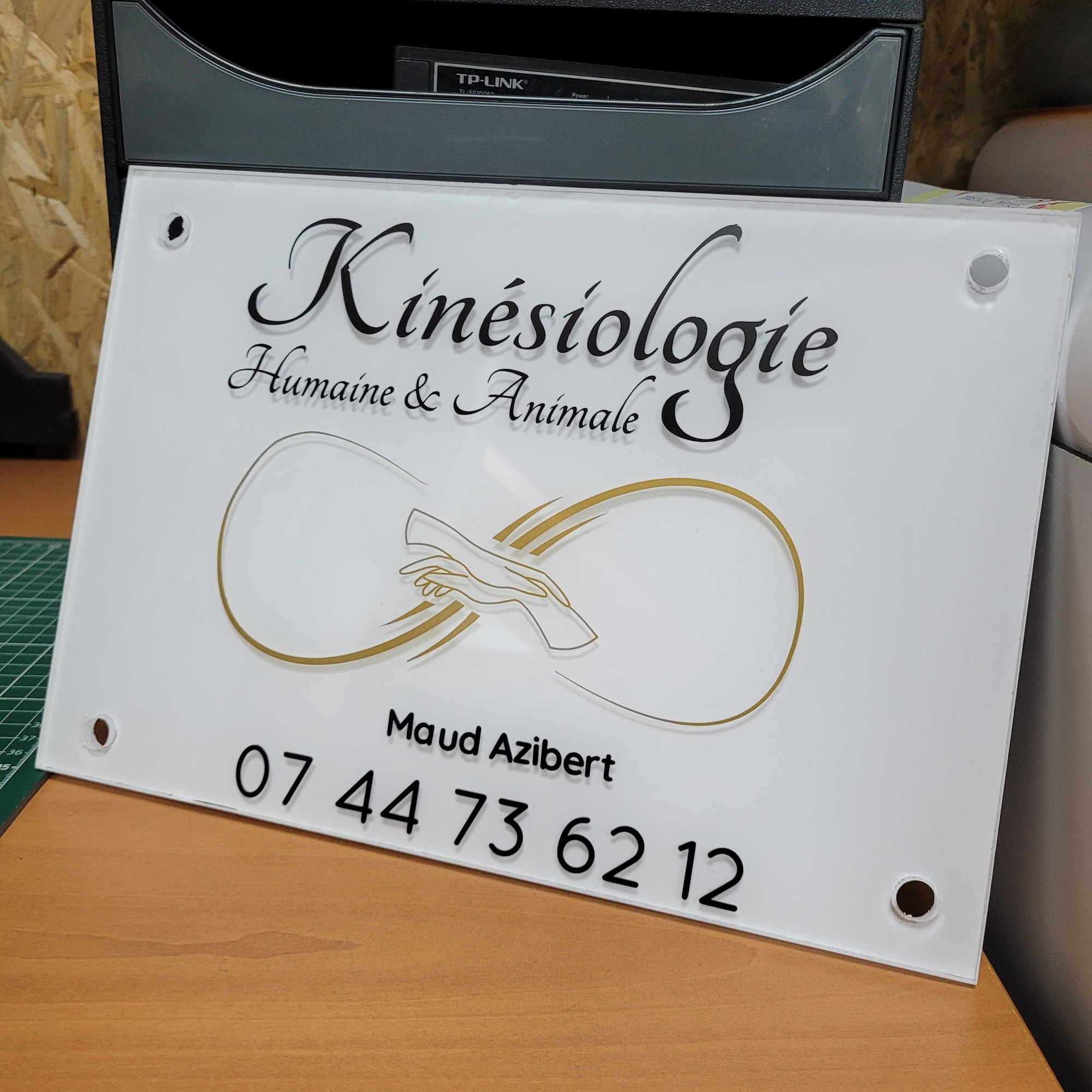 Plaque professionnelle rectangulaire en plexiglas blanc posée sur un plan de travail d'atelier, percée aux quatre angles pour recevoir des entretoises, portant un logo doré et un numéro de téléphone