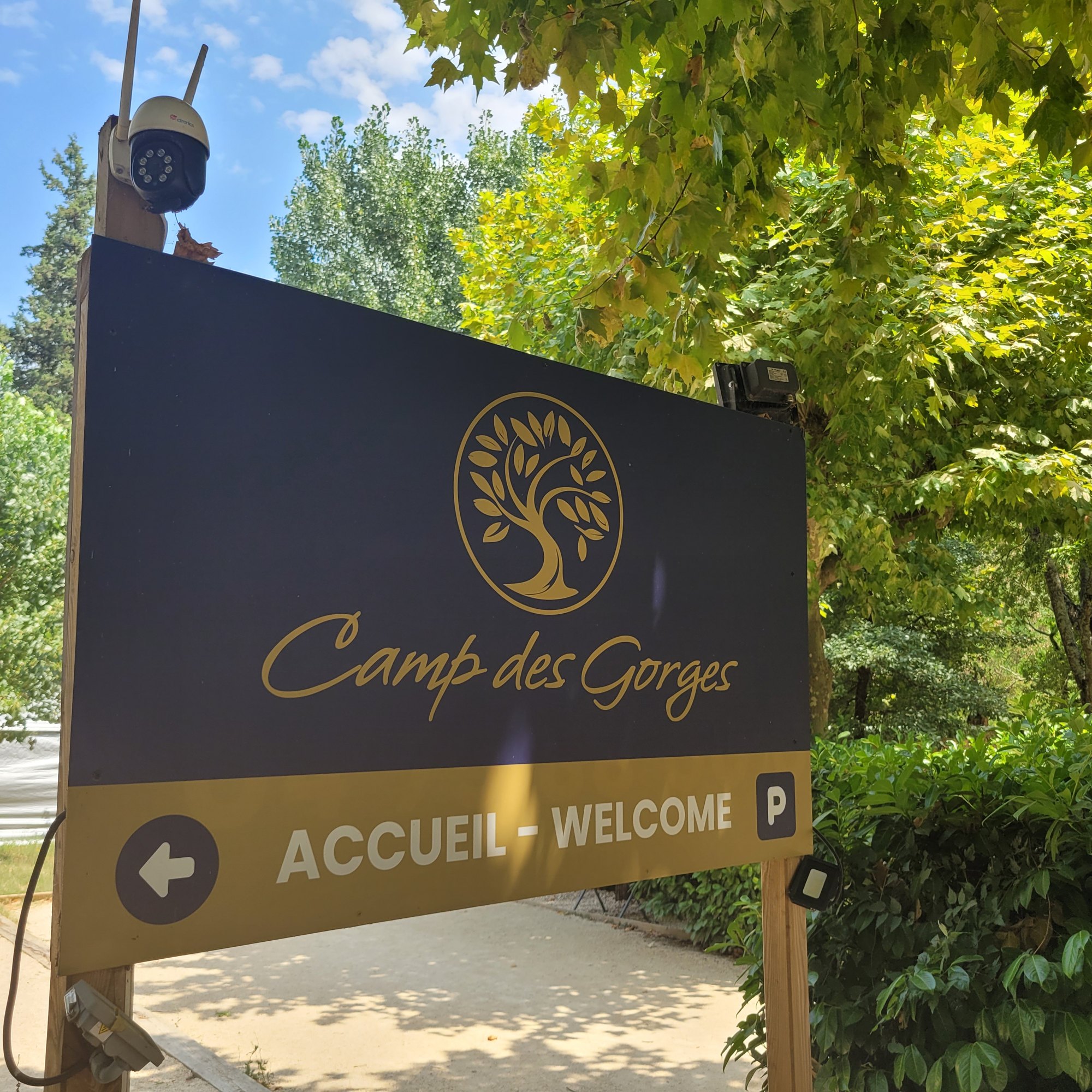 Panneau imprimé bleu nuit et doré monté sur deux poteaux bois à l'entrée d'un camping, portant un logo, le nom de l'établissement et un bandeau directionnel Accueil-Welcome avec flèche et pictogramme de parking