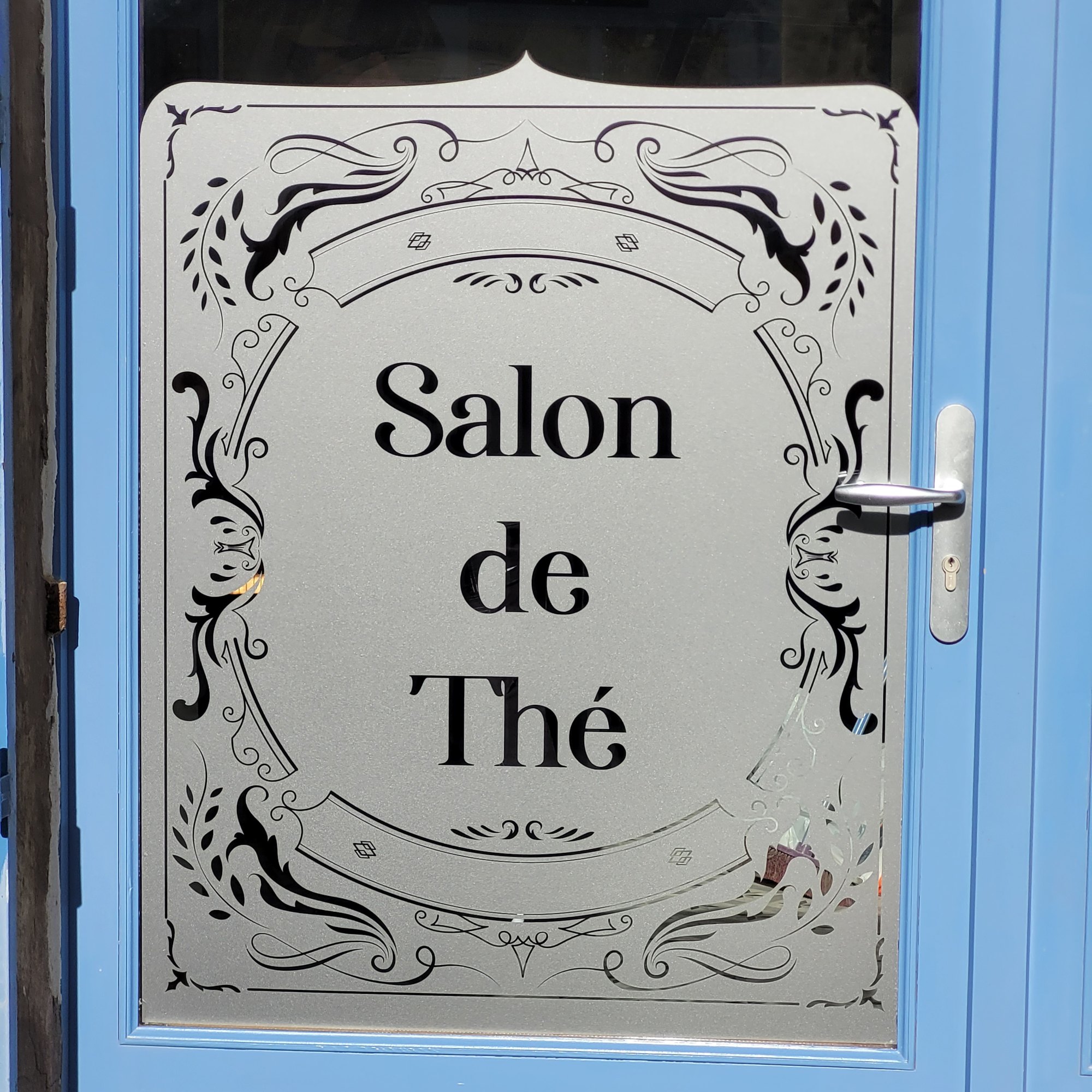 Porte vitrée bleue dont le vitrage est couvert d'un film dépoli découpé en encadrement ornemental, avec les mots Salon de Thé réservés en transparence